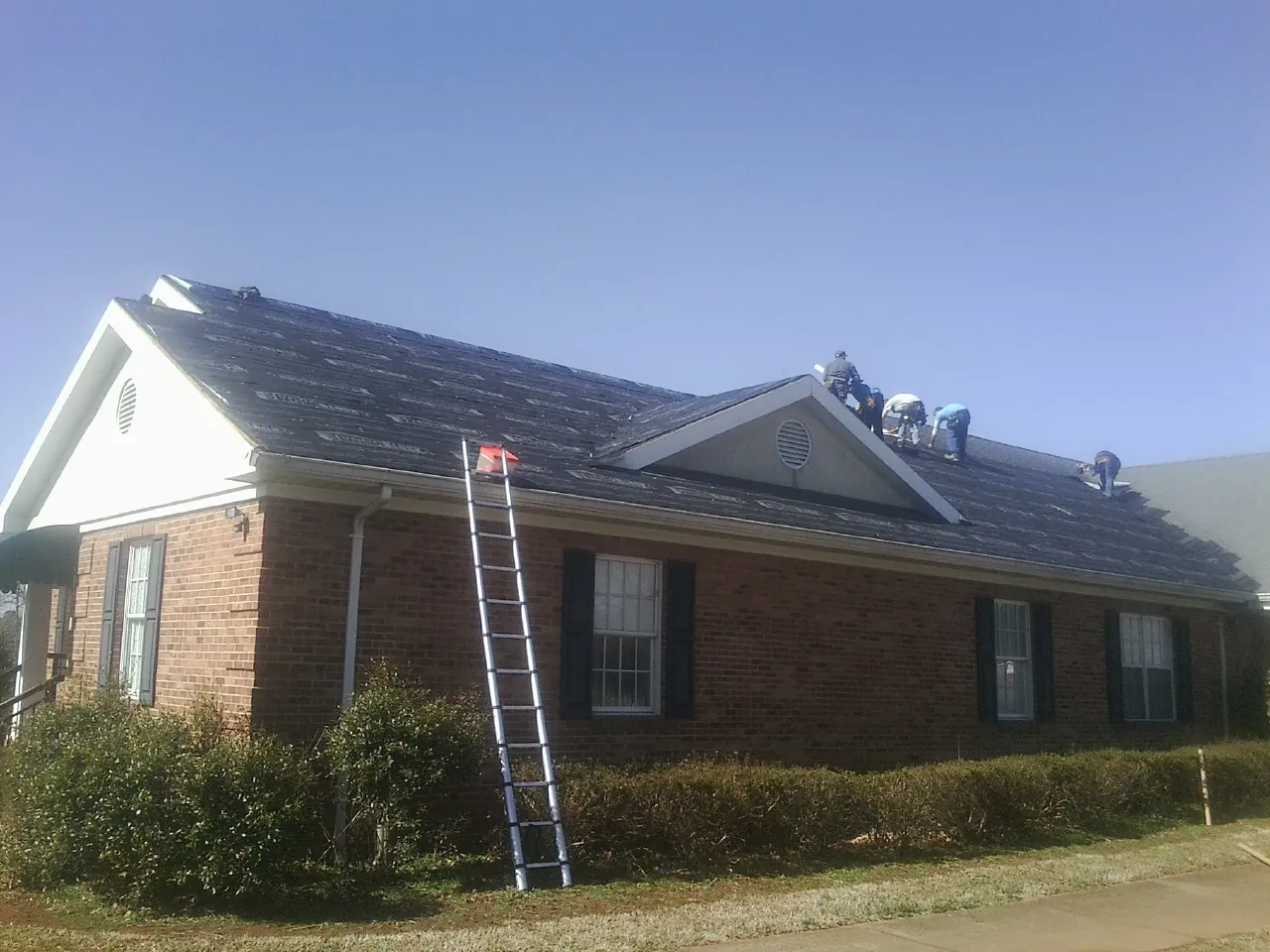 Crew tearing shingles off a brick ranch roof with a ladder set — Metal Roof Repair work by our crew in Columbiaville