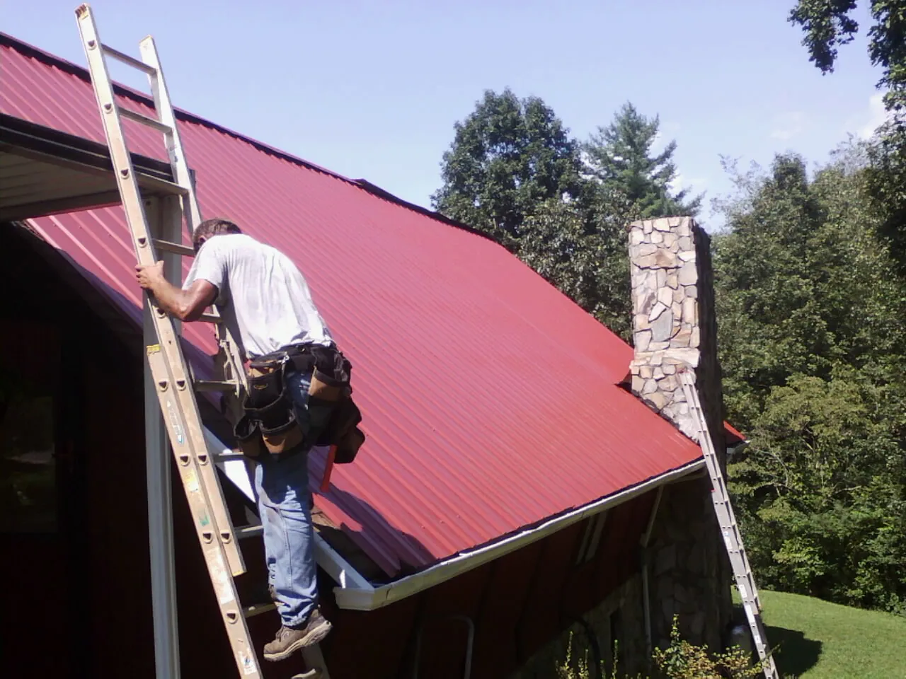 Roofer on a ladder beside a red metal roof and stone chimney — Soffit & Fascia Repair by Roofing Brothers in Columbiaville